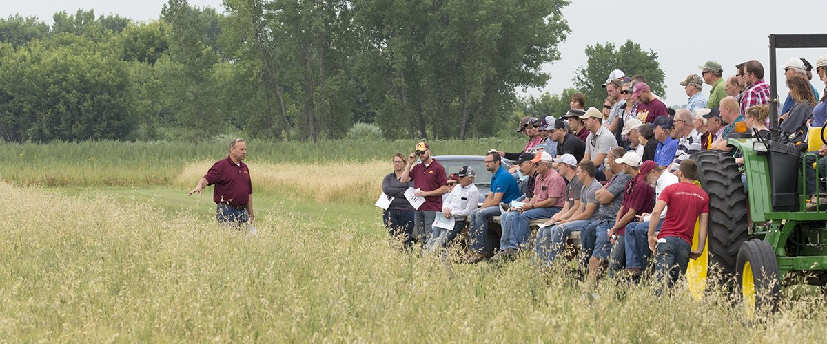 A man stands at the edge of a field and presents to a group of people on hayracks 